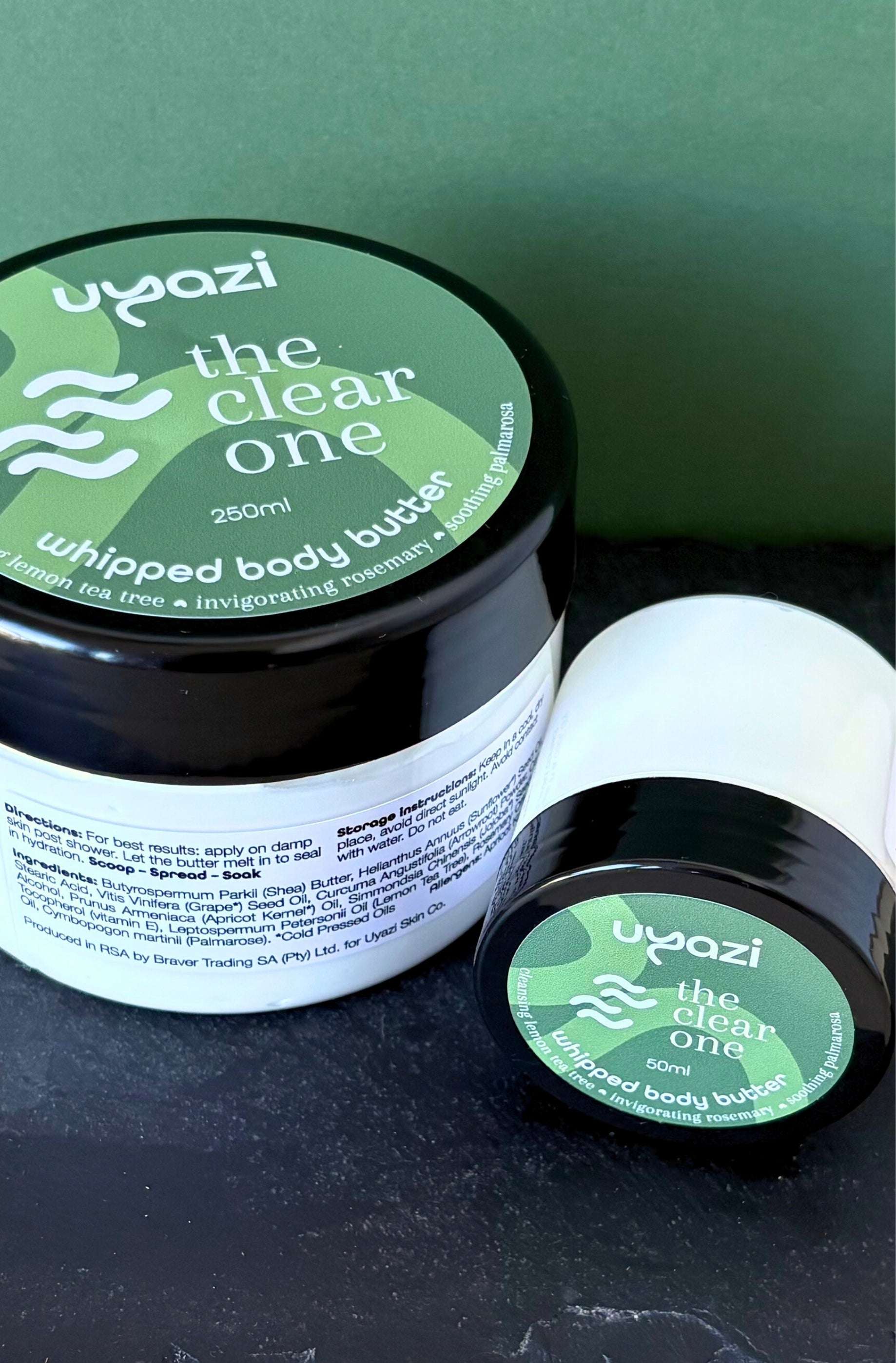 Two containers of 'the clear one' whipped body butter by uyazi on a dark surface with a green background.