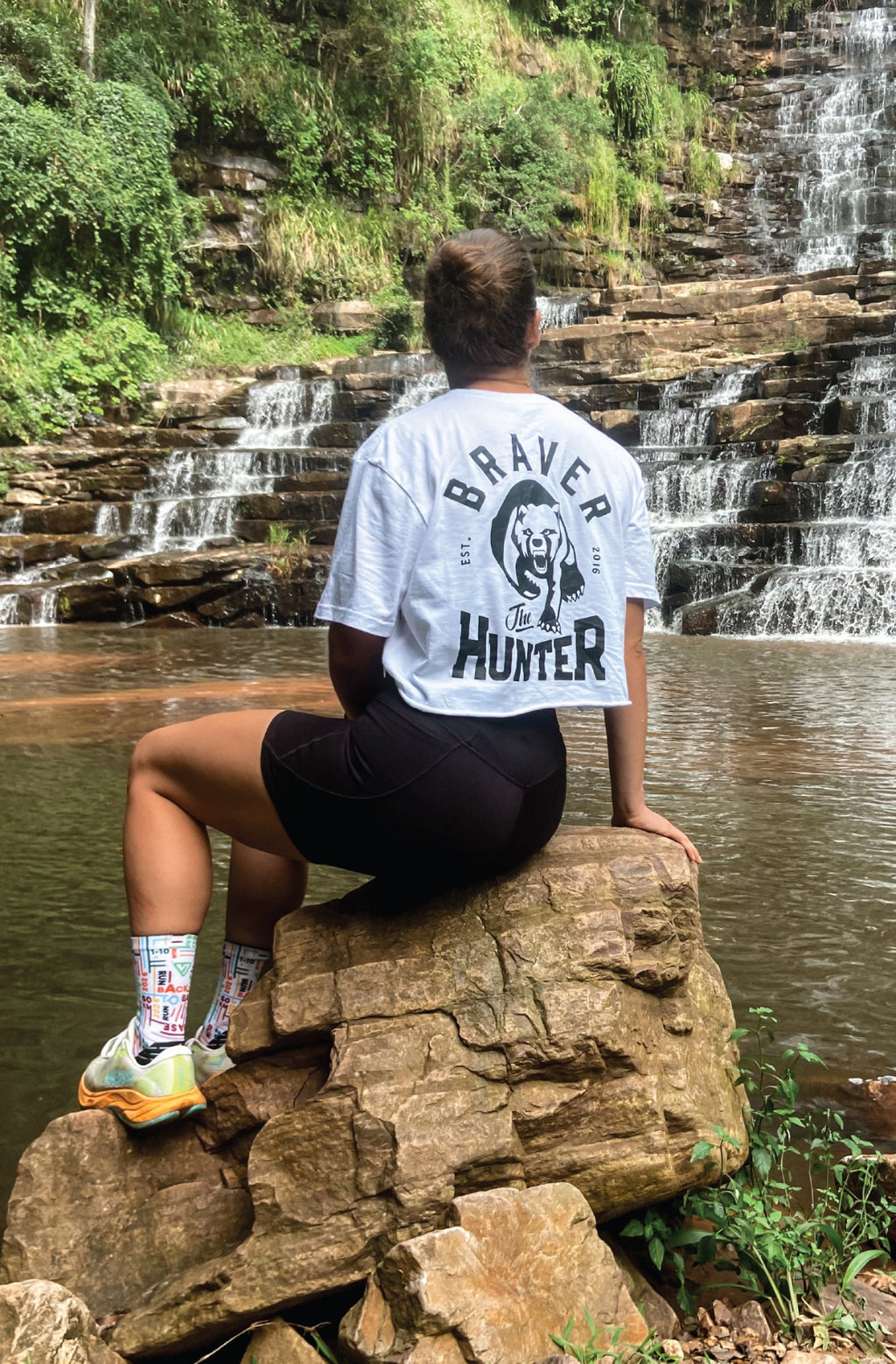 Woman sitting on a rock by a waterfall wearing a 'Braver Hunter' white graphic cropped t-shirt.