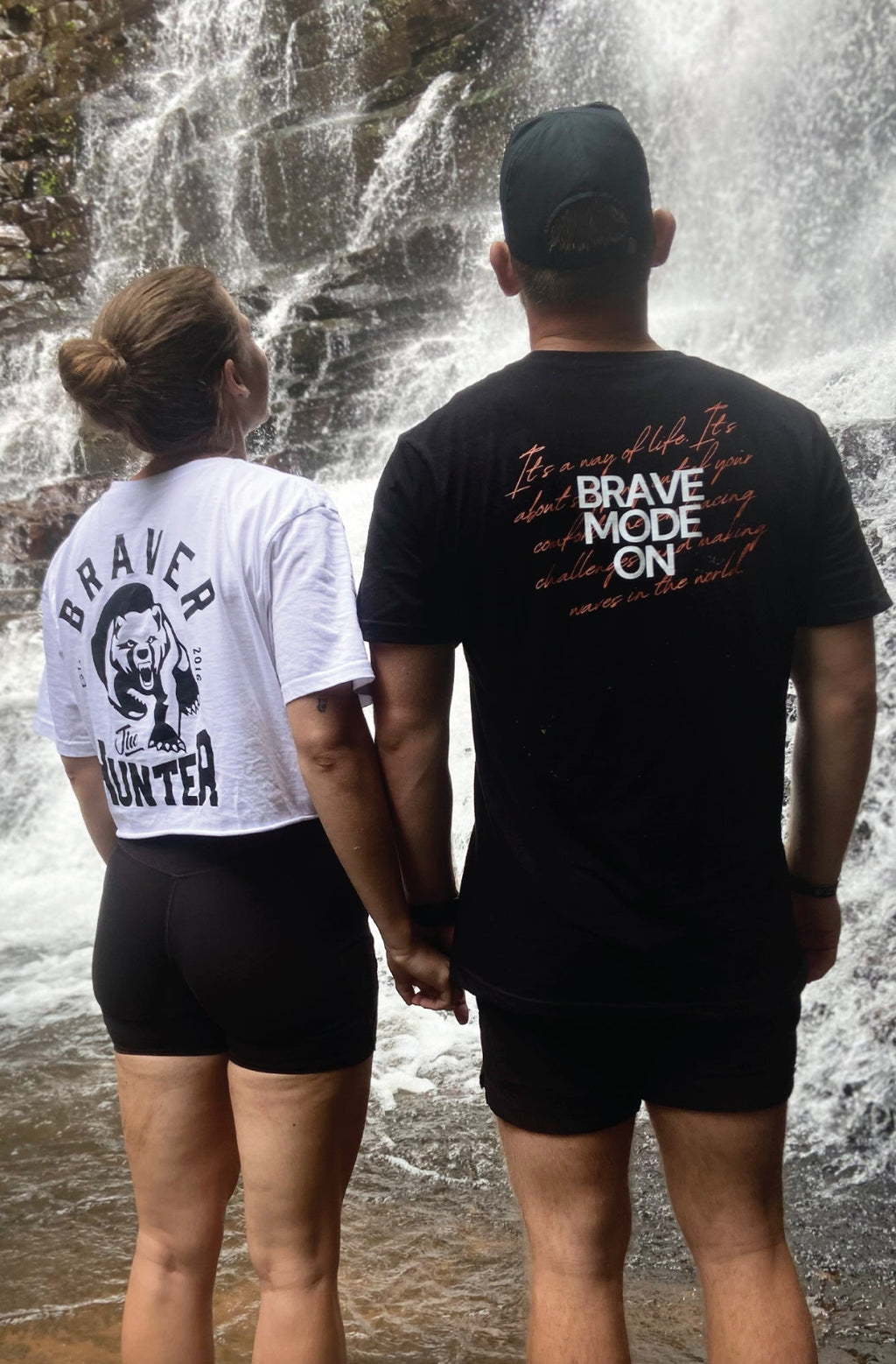 Two people standing in front of a waterfall wearing braver t-shirts with text and graphics.