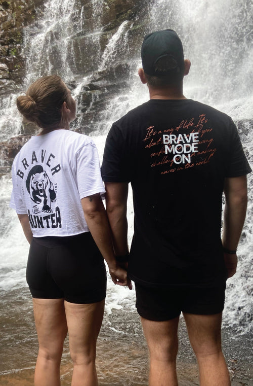 Two people standing in front of a waterfall wearing braver t-shirts with text and graphics.