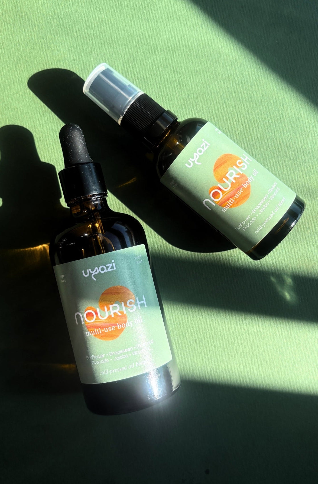 Two bottles labeled 'Nourish' multi use body oil on a green surface with shadows.